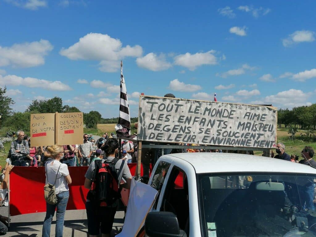 Photographie d'une manifestation organisée par la CAMIL en 2021 contre l’installation d’une usine d’enrobé à Puceul sur le site d’implantation du projet. Sur une pancarte on peut lire "tout le monde aime les enfants mais peu de gens se soucient de leur environnement."